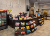 Café with snack racks, assorted chips, candies, a soda fridge, and a bakery display. Modern lighting and decor with sunflower in a vase on the counter.