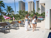 Four people walk by a poolside area with lounge chairs, palm trees, and tall buildings in the background on a sunny day.