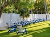 Rows of blue and white Adirondack chairs are arranged on green grass near a white fence, with trees and string lights overhead.