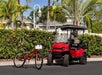 A red bicycle and a red golf cart are parked side by side on a paved road, with tropical plants and white houses in the background.