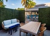 Outdoor patio area with a sofa, two small blue tables, a wooden dining table with wicker chairs, hedge walls, and a view of a poolside bar and palm trees in the background.