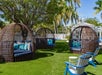 Three round wicker cabanas with blue cushions sit on a green lawn, surrounded by palm trees and white Adirondack chairs under sunny skies.
