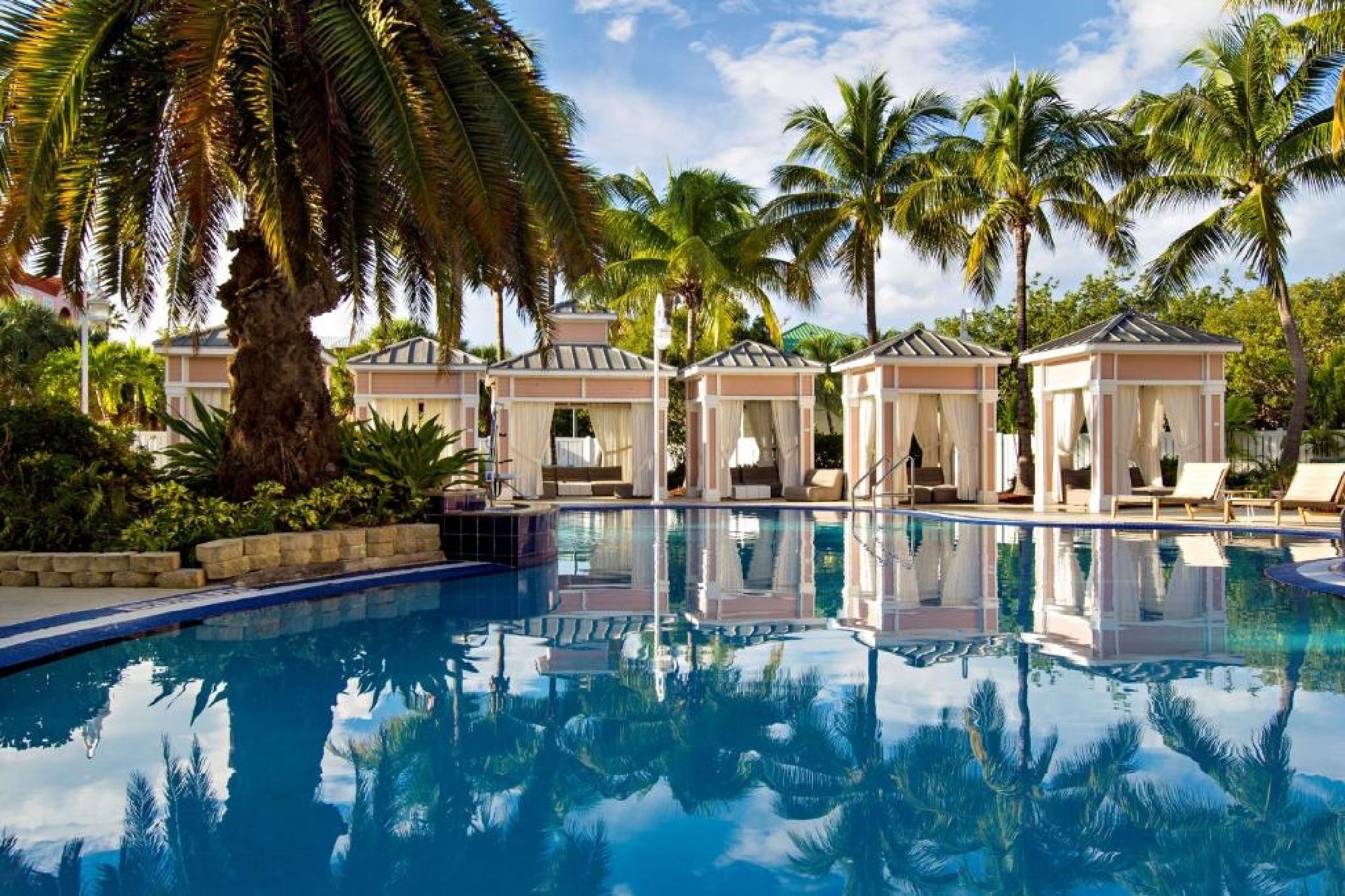 Outdoor swimming pool with clear blue water, surrounded by palm trees and poolside cabanas under a partly cloudy sky.