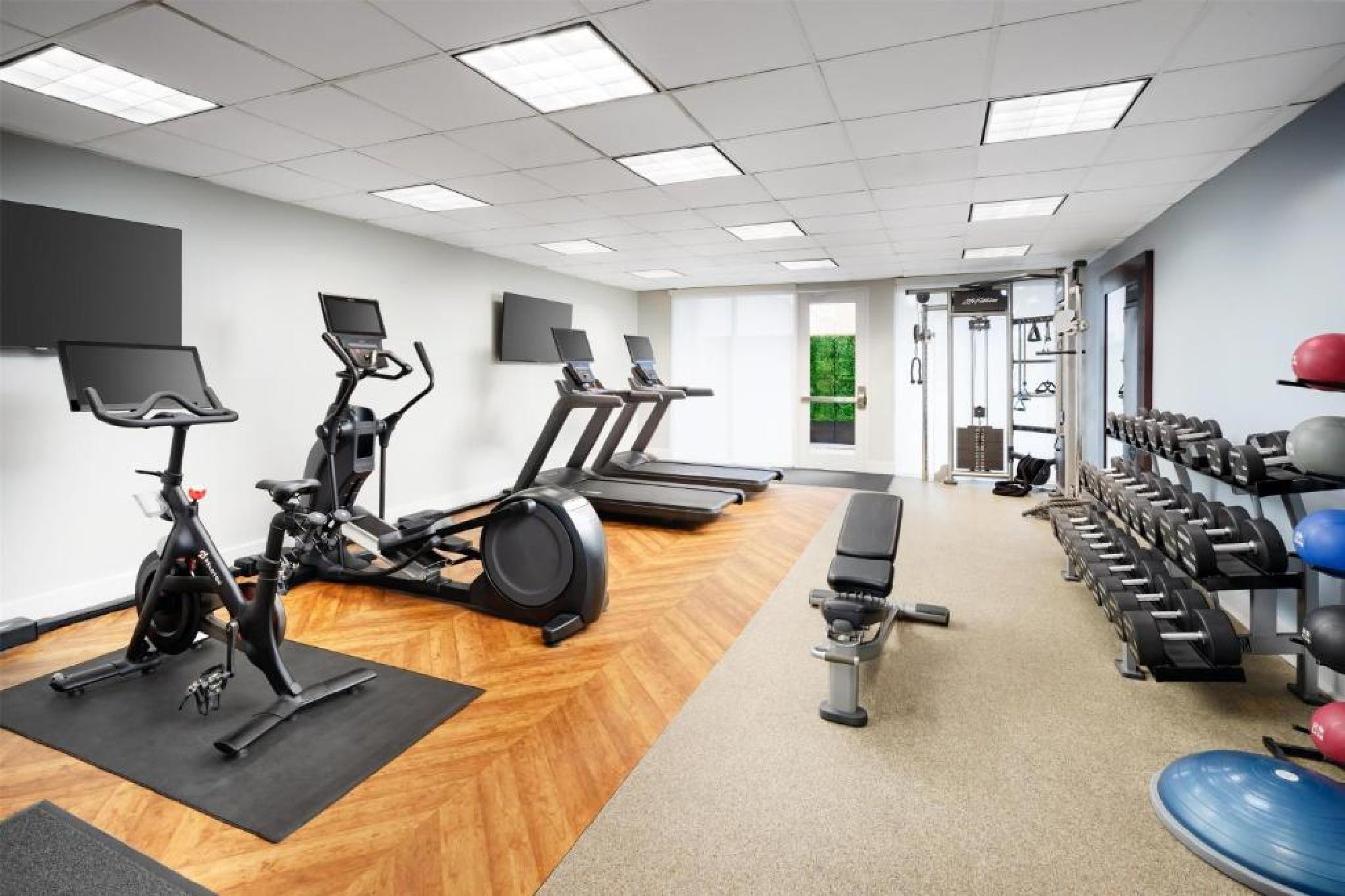Modern gym with treadmills, stationary bike, elliptical machine, weight bench, dumbbells, exercise balls, and cable machine on wood and carpeted flooring.