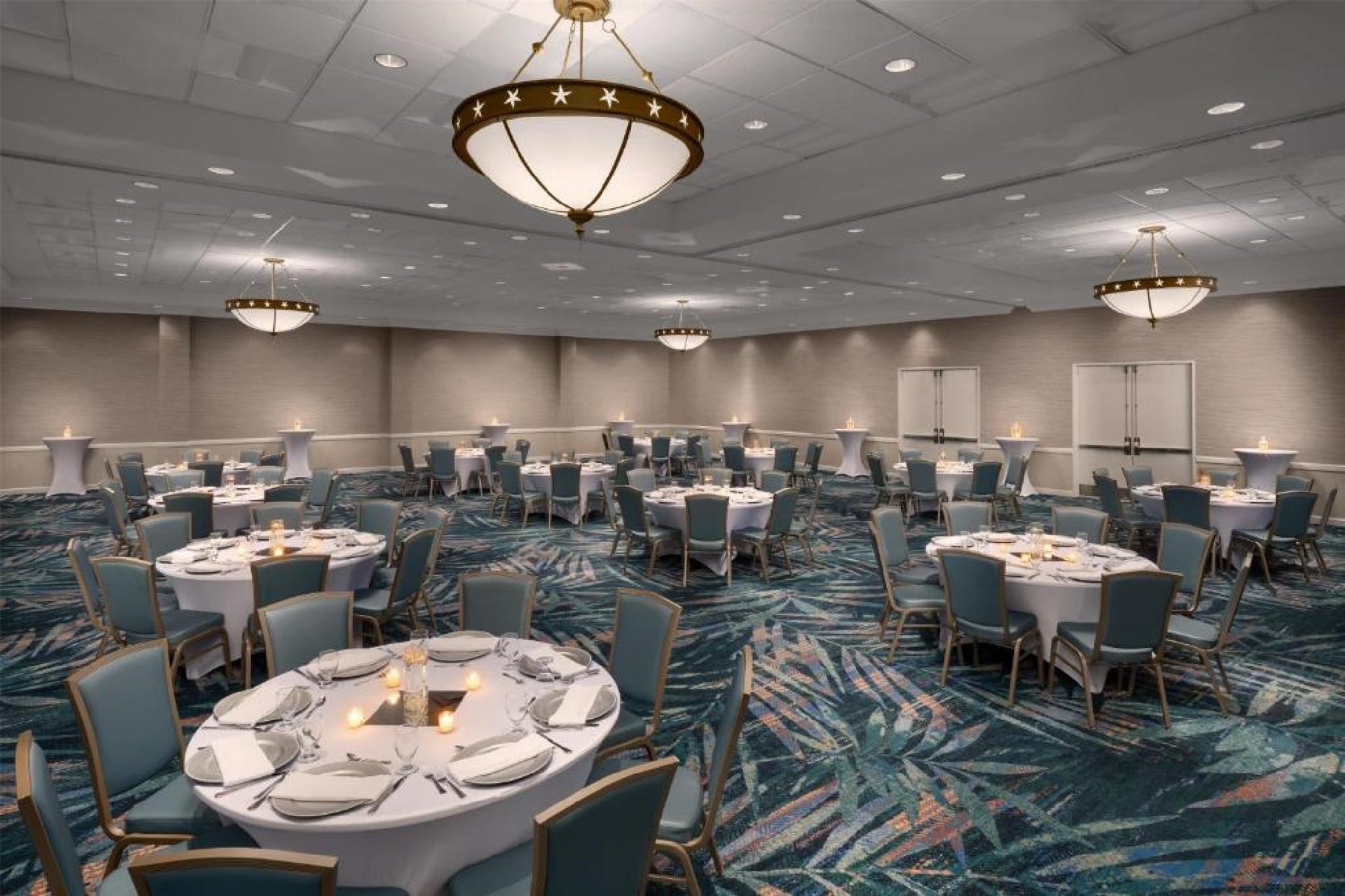 Large banquet hall with round tables set for dining, blue chairs, lit candles, patterned carpet, and modern chandeliers.