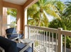 A balcony with two blue cushioned chairs and a small table overlooks palm trees and a sunny outdoor area.