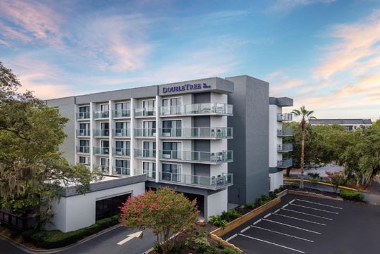 A five-story hotel building with a modern design, featuring balconies and surrounded by trees. The parking area is partially visible in the foreground under a clear sky.