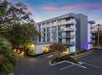 Exterior of a modern hotel with a gray facade, multiple balconies, and a parking lot in front, set against a colorful sunset sky. Lush greenery surrounds the building.