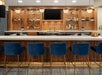 A modern bar with blue stools, a wooden counter, and shelves displaying various bottles. A large TV screen is mounted above the counter.