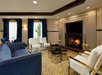 A cozy living room with a lit fireplace, blue sofa, white armchairs, a glass coffee table, and large windows with dark blue curtains.