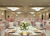 Elegant banquet hall with round tables set for an event. Each table has white tablecloths, pink sashes, floral centerpieces, and dinnerware. Chairs are draped in white with pink bows.