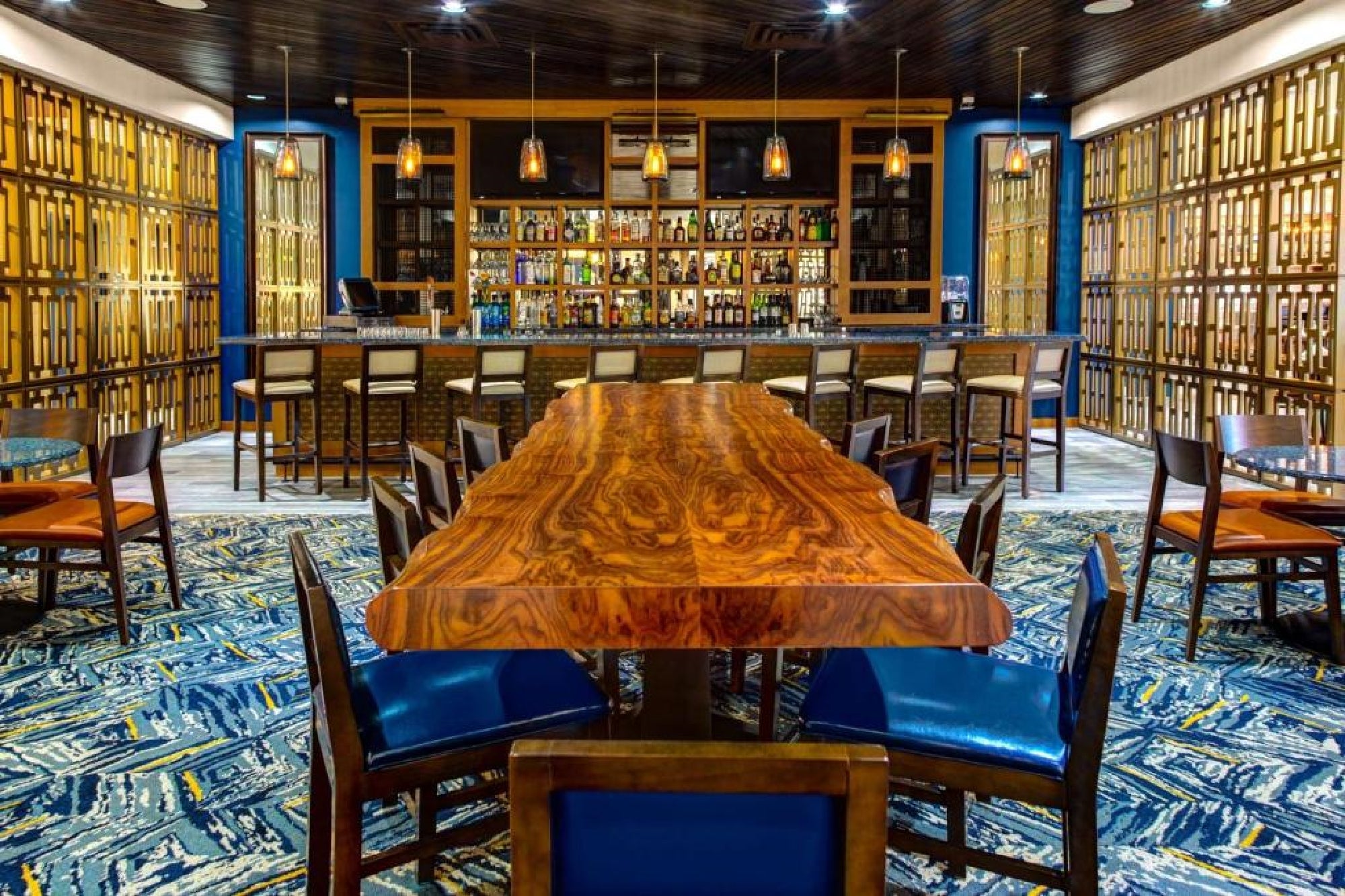 A long wooden table with chairs is centered in a modern bar with shelves of liquor bottles, pendant lights, and patterned blue carpet and walls.