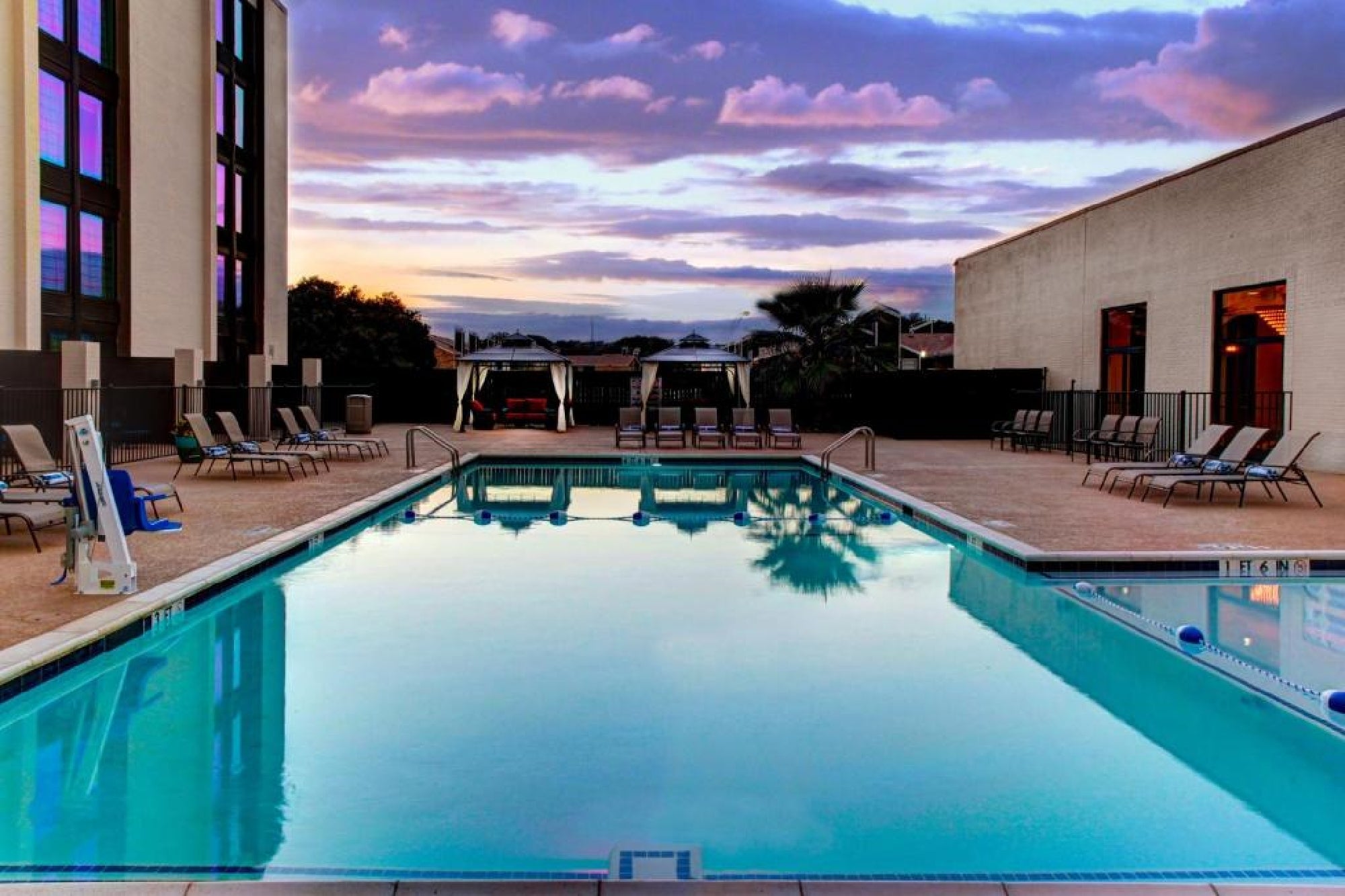 Outdoor swimming pool surrounded by lounge chairs, with a building on each side, at sunset under a partly cloudy sky.