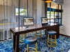 Two desktop computers with keyboards and mice are set up on a wooden table with three barstools in a hotel business center area with patterned carpet and floor-to-ceiling curtains.
