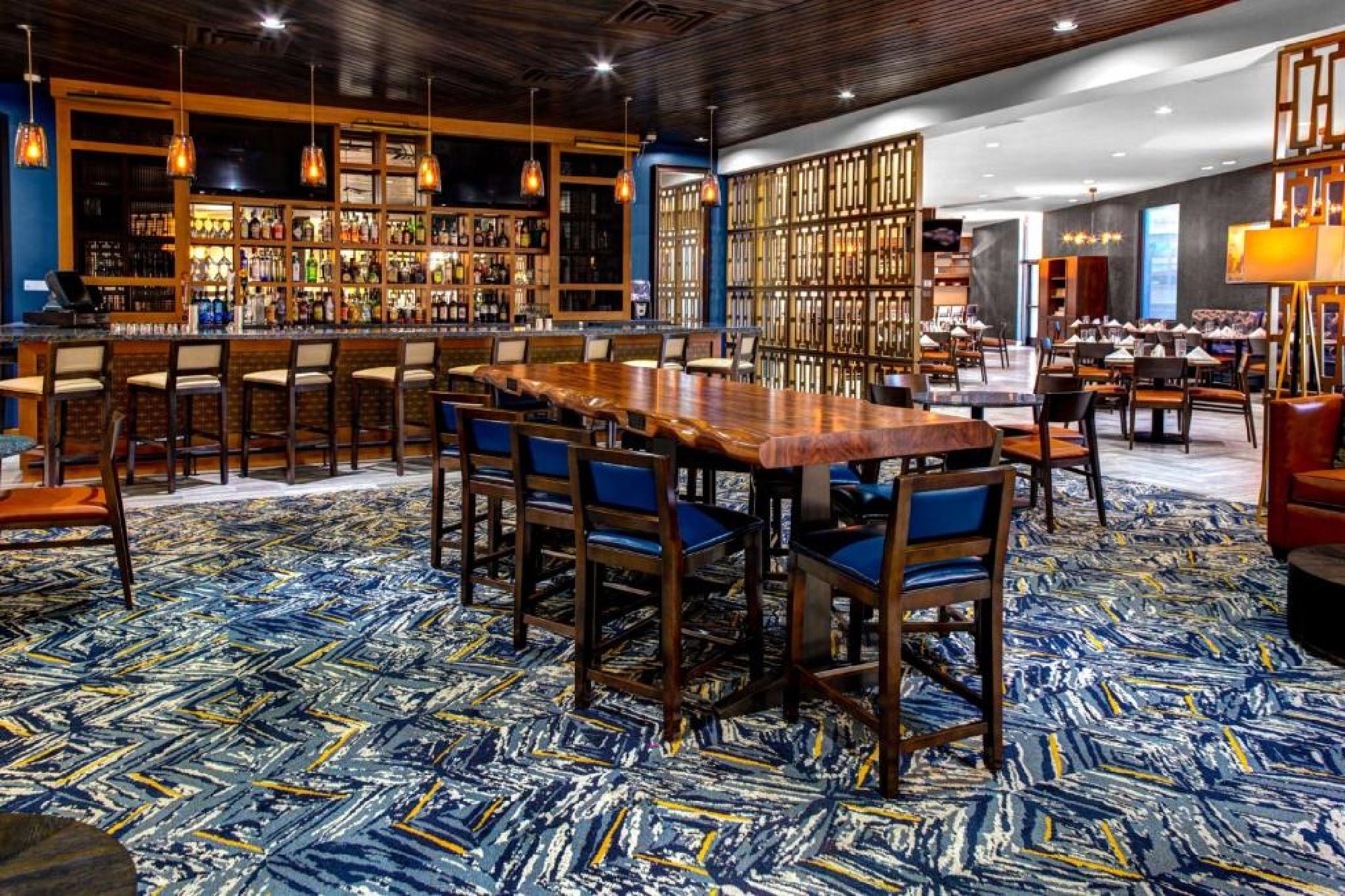 Modern hotel bar with a long wooden communal table, high-backed chairs, patterned carpet, and shelves of bottles behind the bar counter. Warm lighting and contemporary decor.