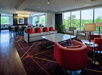 Modern lounge area with large windows, a sofa set with red cushions, a glass coffee table, red armchairs, a dark carpet with geometric patterns, and a dining table in the background.