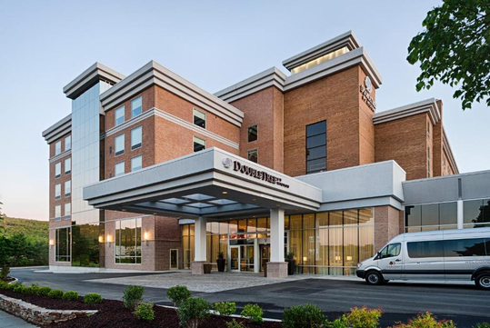 A modern, multi-story hotel building with large windows and a canopied entrance. A shuttle van is parked in front.