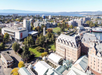 Aerial view of a cityscape featuring historic and modern buildings, green parks, and a waterfront with distant mountains under a clear blue sky.