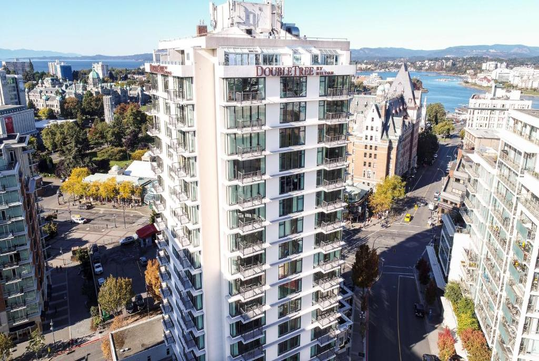 Aerial view of a tall white hotel building in an urban area with trees and streets, near a body of water and distant hills.