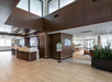 A modern hotel lobby with wooden accents, a reception desk, seating area, and a plant. A sign on a stand is near the desk.