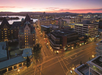 Aerial view of a city intersection at dusk with buildings, illuminated streets, and a visible waterfront in the background under a colorful sky.