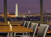 A restaurant interior with wooden tables and gray chairs overlooks the Washington Monument and U.S. Capitol at dusk through large windows.