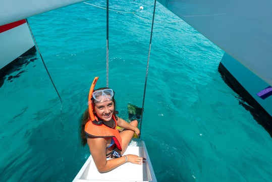 A person wearing a snorkel and life vest sits on a boat ladder above clear turquoise water.