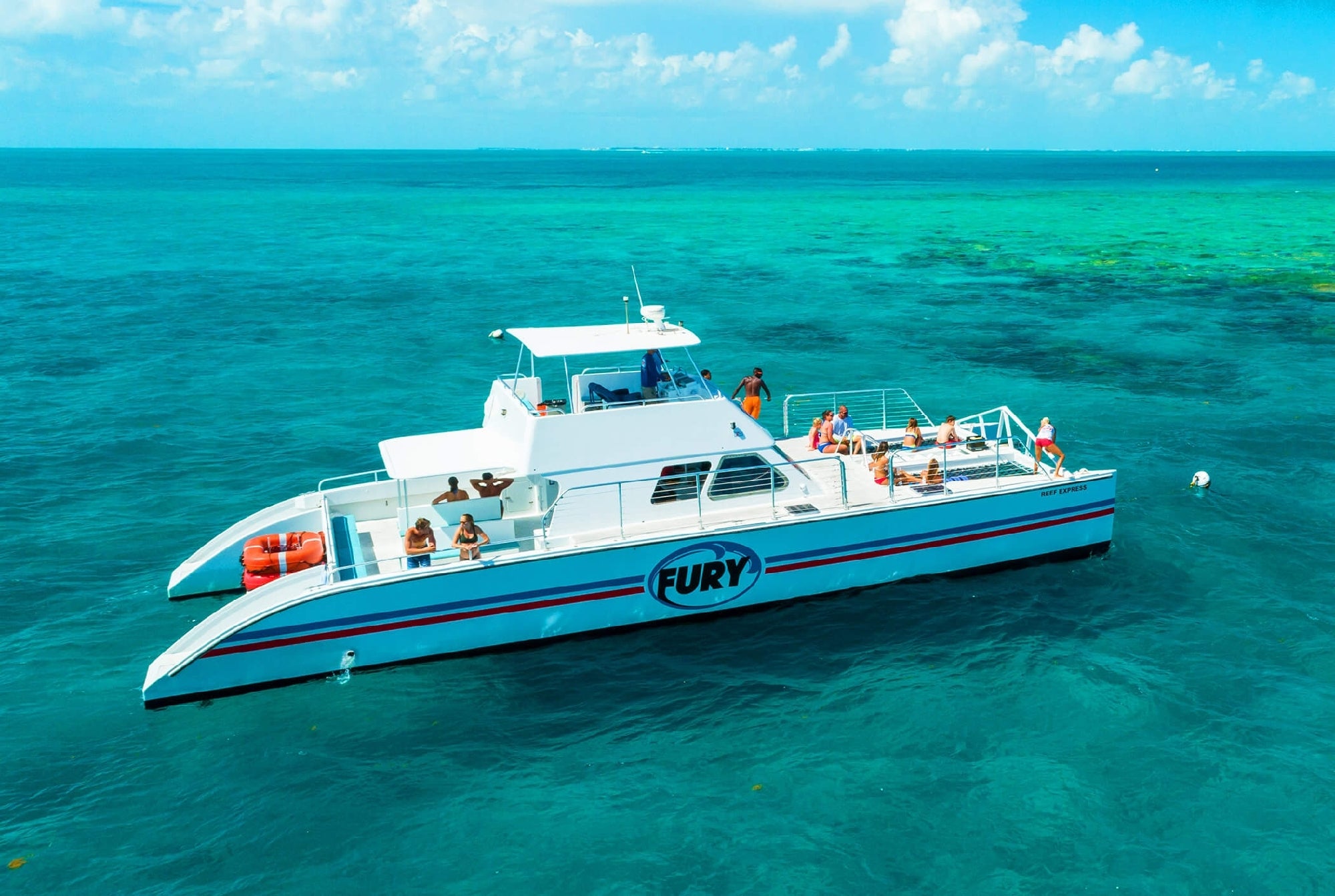 A white catamaran named "Fury" is anchored in clear turquoise waters with passengers on board under a partly cloudy sky.
