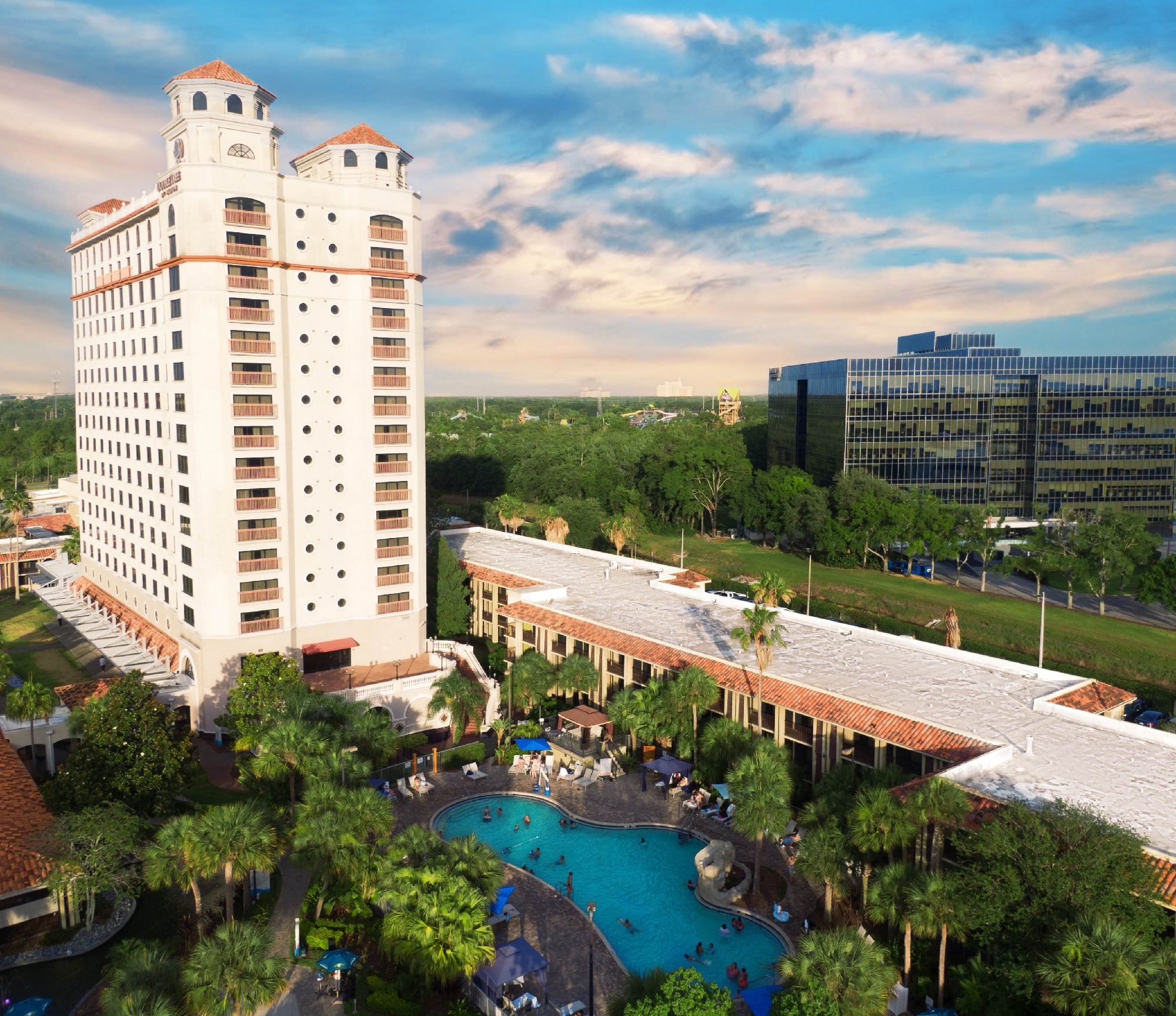 A tall hotel tower with balconies stands next to a large outdoor pool surrounded by palm trees, with office buildings and greenery in the background under a partly cloudy sky.