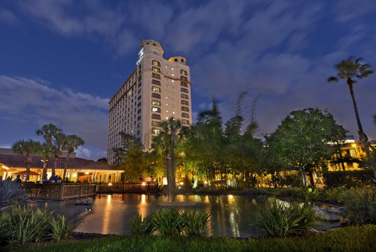 Tall hotel building with illuminated windows at dusk, surrounded by palm trees, landscaped gardens, and a pond with a small fountain in the foreground.
