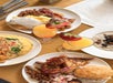 Four plates of breakfast foods including omelet, biscuits and gravy, bacon, sausage, French toast, hash browns, oatmeal with raisins, and two orange juice glasses garnished with strawberries.