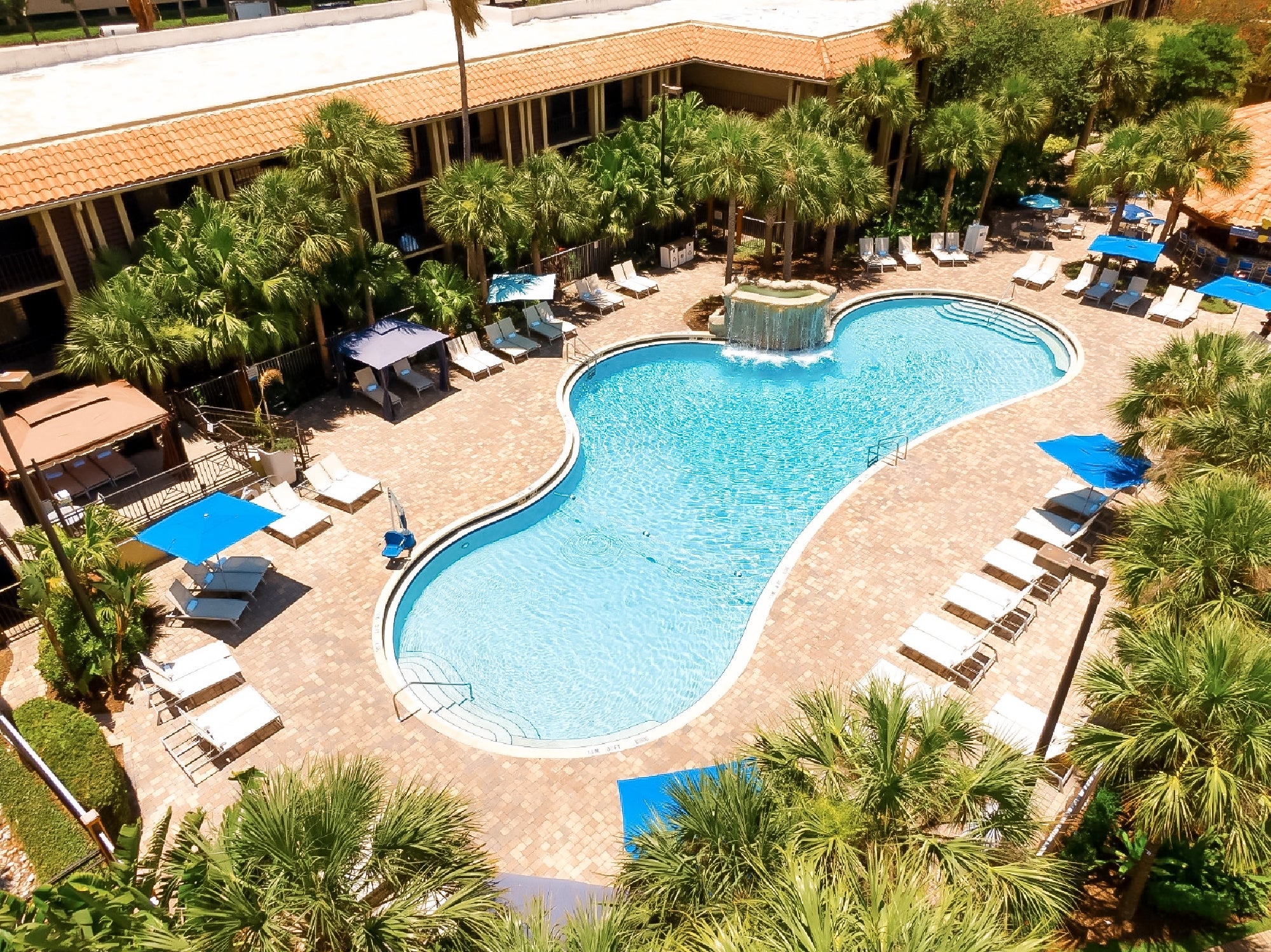 Aerial view of a hotel courtyard featuring a large kidney-shaped swimming pool, lounge chairs, palm trees, cabanas, blue umbrellas, and a small waterfall feature.