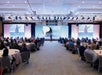 A speaker presents on stage to an audience seated at round tables in a large conference room with projection screens and lighting equipment.