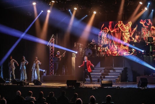A man in a red suit jacket standing center stage with three people to the left, a band behind him, and strobe lights around the room.