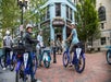 People on rental bikes pause near a blue building labeled "French Broad Chocolate Lounge." Tree-lined street with pedestrians in the background.
