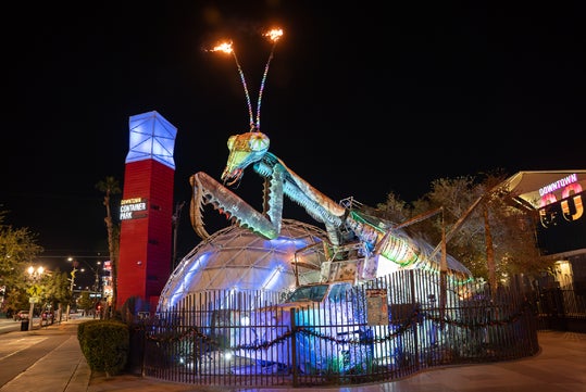 A large, illuminated praying mantis sculpture with flaming antennae stands outside a geodesic dome at Downtown Container Park at night.
