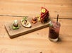 A wooden board with assorted appetizers, including a meatball, bread with toppings, bacon-wrapped item, and layered sandwich, next to a pink cocktail on a wooden table.