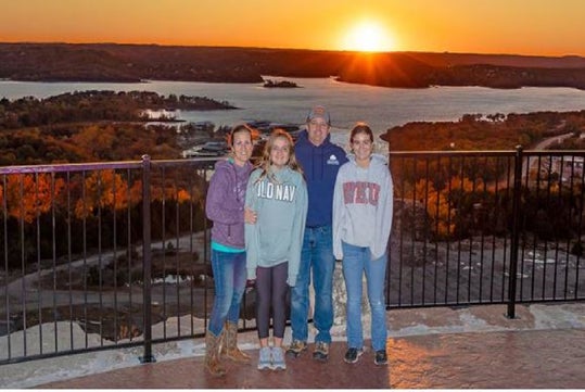 A family of four stands on a scenic overlook at sunset, with a view of a lake and hills in the background.