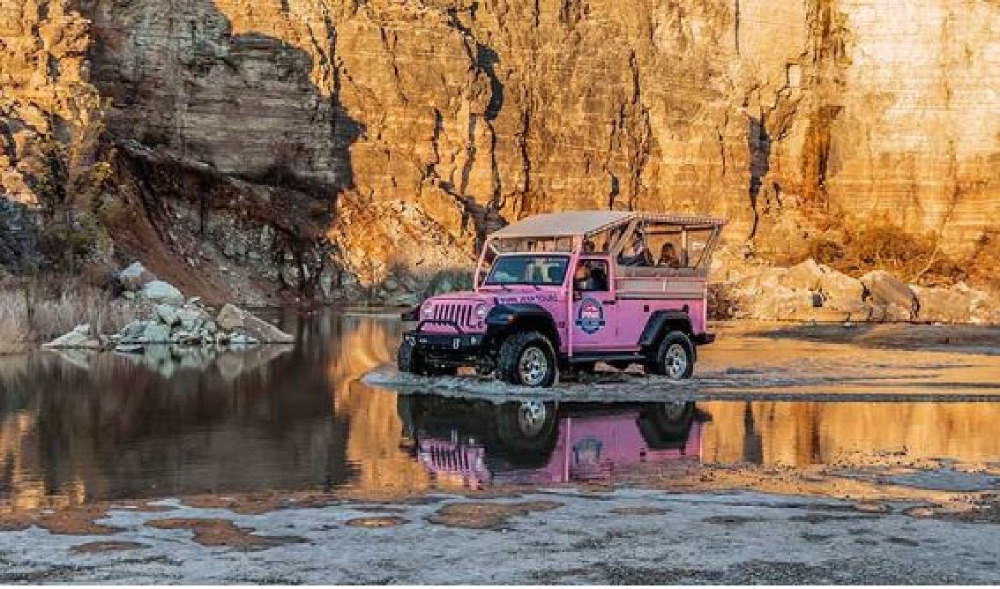 A pink jeep drives through shallow water with a rocky cliff in the background.