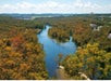 Aerial view of a winding river surrounded by dense forest with autumn foliage, under a partly cloudy sky. Buildings are visible in the distance.