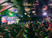 A performer on stage at a vibrant drag show venue. An audience sits at tables under colorful lights and chandeliers.