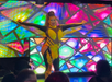 Performer on stage in a yellow and black outfit with fringes, arms outstretched. Colorful geometric backdrop and audience in foreground.