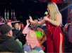 A woman in a red jumpsuit stands with a microphone, playfully interacting with an audience member in a lively setting.
