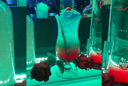 A colorful layered cocktail with whipped cream and a cherry on top, surrounded by red roses, against a backdrop of bottles in neon lighting.