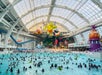A large indoor water park with a wave pool filled with people. Overhead, there's a glass ceiling and giant floating inflatable characters. A long water slide is visible on the left.