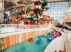 Indoor water park with a lazy river, colorful water slides, and play structures. People are floating in tubes under a large, glass-paneled roof.