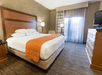 Hotel room with a large bed, orange throw, wooden furniture, lamp, and a window with sheer curtains. A flat-screen TV sits on a dresser. Neutral-toned walls and carpet.