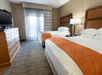 Hotel room with two double beds, orange accents, a flat-screen TV, and a dresser. A floor lamp sits between the beds, and a window with sheer curtains is in the background.