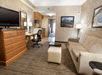 Hotel room with a couch, desk and chair, TV on a dresser, and a small kitchen area in the background. Walls have framed art, and there are two lamps. Carpeted floor with a footstool.