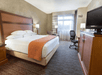 Hotel room with a large bed, an orange throw, two lamps, a window with curtains, a desk, a chair, and a TV.